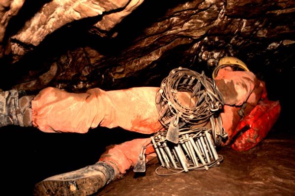 Explorer hauling wire ladder equipment through muddy low passage in Rat's Nest Cave, Canmore Alberta — early exploration of Canadian Rockies cave system in the 1970s
